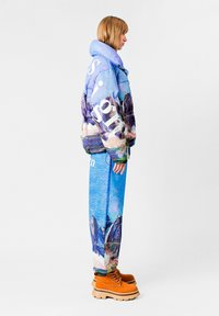 Chaqueta acolchada con estampado de montañas en azul y púrpura, con texto grande en blanco en un costado. Combinada con pantalones azules texturizados y botas marrones con cordones.