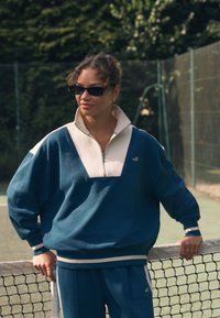 Felpa con cappuccio con zip blu e crema caratterizzata da un colletto alto e una silhouette oversize, abbinata a pantaloni blu coordinati. Accentuata da un logo.