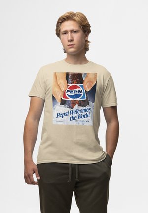 Giovane uomo che indossa una maglietta beige con una bottiglia vintage di Pepsi e la scritta "Pepsi Welcomes the World" sul davanti.