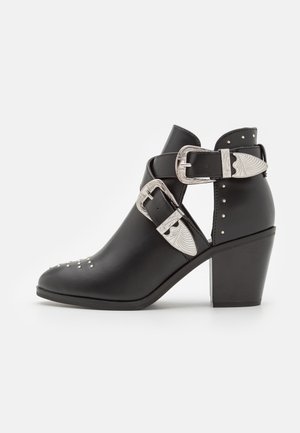 Bottine noire à talon épais, boucles décoratives argentées, découpes latérales et petits clous argentés sur la zone des orteils et du talon.