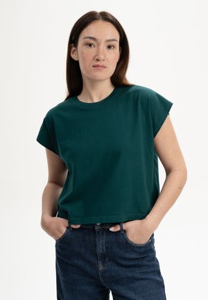 Vrouw met lang bruin haar, gekleed in een donkergroene top met korte mouwen en donkerblauwe spijkerbroek, staat met handen in de zakken tegen een witte achtergrond.