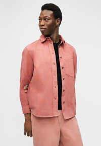 Άνδρας με κοραλλί πουκάμισο με λουλουδάτο μοτίβο και παντελόνι που ταιριάζουν, κοιτάζει πάνω από τον ώμο με το ένα χέρι πίσω από την πλάτη σε απλό φόντο.