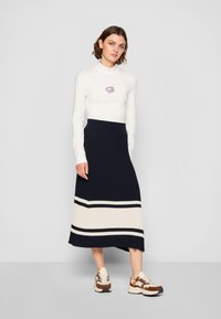 Hvid langærmet top med logo, parret med en marineblå ribbet nederdel med creme- og marineblå striber, samt beige-brune sneakers med sorte accenter.