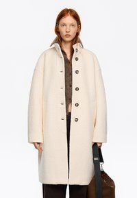 Mujer joven de cabello rojo que lleva un abrigo crema extragrande con botones, suéter marrón, pantalones negros y sostiene una bolsa marrón con una correa negra.