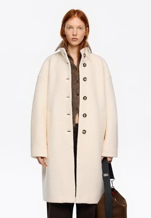 Mujer joven de cabello rojo que lleva un abrigo crema extragrande con botones, suéter marrón, pantalones negros y sostiene una bolsa marrón con una correa negra.