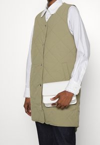 Gequilted olijfgroene vest met knoopsluiting, gedragen over een witte blouse met golvende manchetten. Een witte clutchtas wordt in de hand vastgehouden.