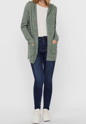 Hellgrüner, lang geschnittener Cardigan mit Kapuze, vorderen Taschen und einer strukturierten Stoffoberfläche. Getragen über einem weißen T-Shirt und dunkelblauen Jeans.