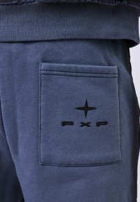 Primer plano de un bolsillo de pantalones de chándal azul marino con un logo de estrella negra y el texto "pxp" debajo de la cinturilla acanalada.