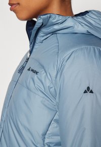 Jasnoniebieska ocieplana kurtka z kapturem, zapinana na zamek, z czarnym logo Vaude. Gładka tekstura z pikowanymi detalami na rękawach.