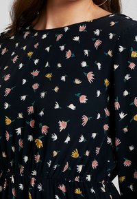 Schwarzes Stoffkleid mit einem floralen Muster in Pink, Weiß, Gelb und Grün. Das Design umfasst eine geraffte Taille und einen runden Ausschnitt.