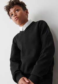 Črn pulover v obliki črke V iz mehkega blaga z dolgimi rokavi in rebrastimi manšetami. Pod njim je viden bel srajčni ovratnik.