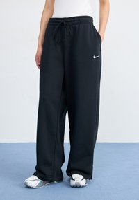 Una persona lleva pantalones de chándal negros con un logo de Nike blanco y zapatillas deportivas blancas, de pie sobre un suelo azul claro frente a un fondo sencillo.