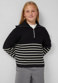 Schwarzer Strickpullover mit einem Reißverschlusskragen, der weiße horizontale Streifen aufweist. Die Ärmel haben ein schwarz-weiß gestreiftes Muster.