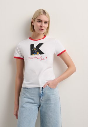 T-shirt blanc à manches courtes avec un piping rouge, portant un "K" noir et un design de personnage de dessin animé. Matière en coton, coupe décontractée.