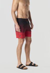 Homme torse nu portant un short de bain dégradé rouge et noir et des sandales noires à double lanière, debout devant un fond clair uni.
