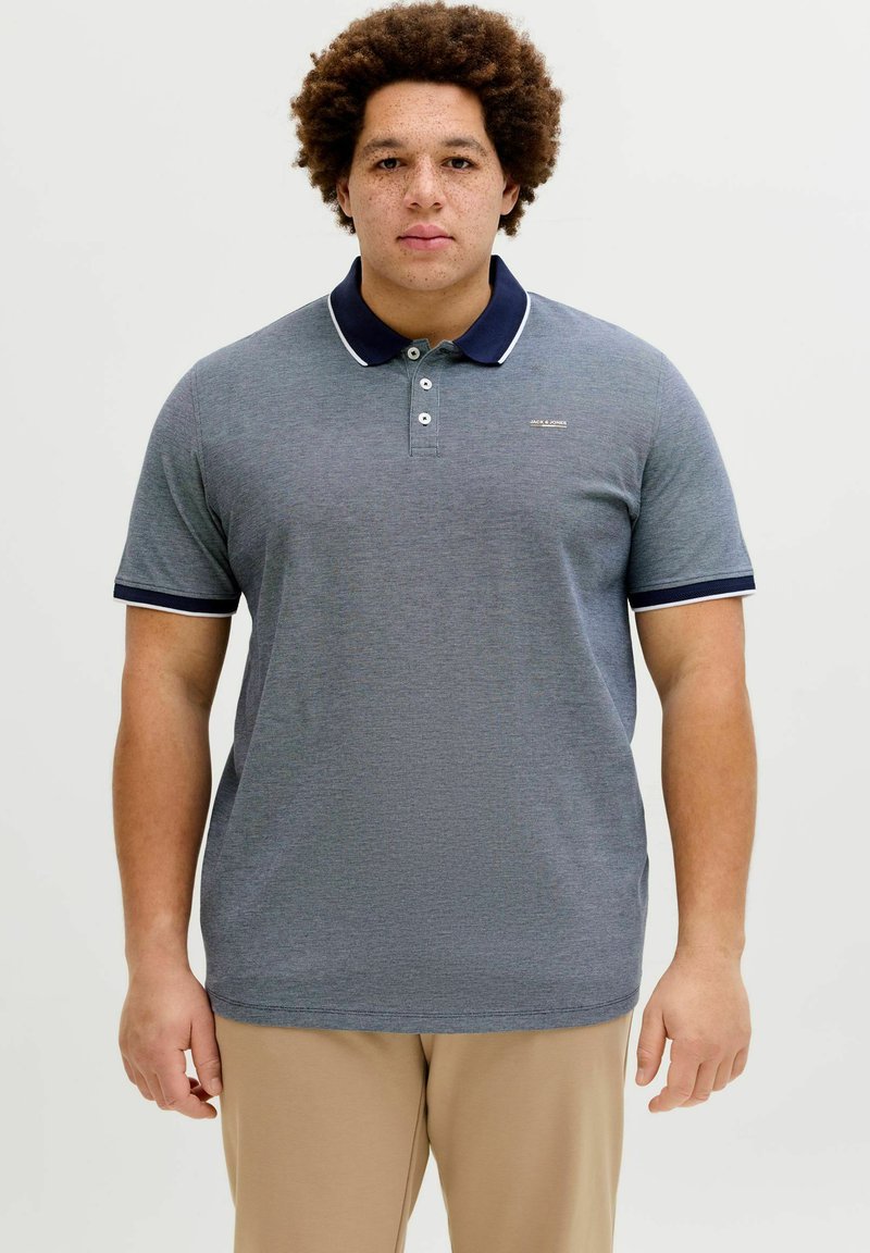 Grå polo med navyblå krave og kant, med tre knapper og tekstureret stof. Båret med beige bukser.