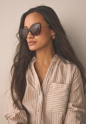 Femme aux longs cheveux noirs portant de grandes lunettes de soleil écaille de tortue, des boucles d'oreilles créoles en or et une chemise beige rayée à boutons.