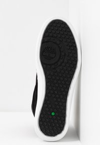 Semelle de chaussure en caoutchouc noir avec un motif de crampons hexagonaux, bord blanc et accent vert au centre. Logo d'arbre embossé.