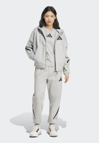 Set sportivo grigio realizzato in materiale morbido, composto da una felpa con zip, una maglietta a girocollo e pantaloni tapered con dettagli neri e logo.