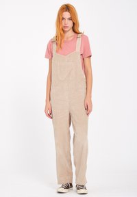 Beige fløjls overaller med en frontlomme, justerbare stropper og en afslappet pasform, parret med en pink T-shirt og sneakers.