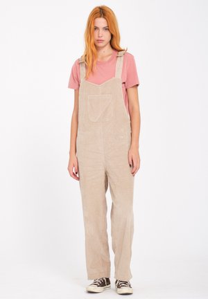 Pettorine in corda beige con tasca frontale, spalline regolabili e vestibilità ampia, abbinate a una maglietta rosa e sneakers.