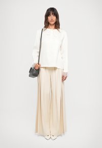 Hvit bluse med blonde mansjetter og snøring i halsen, sammen med vide beige bukser og en liten grå veske. Glatt stoff, enkelt design.