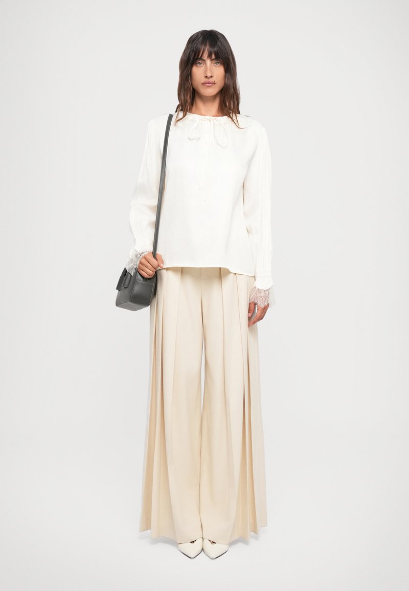 Hvit bluse med blonde mansjetter og snøring i halsen, sammen med vide beige bukser og en liten grå veske. Glatt stoff, enkelt design.