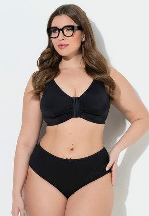 Kvinde med langt hår og briller iført en sort bh med lukning foran og matchende trusser med høj talje, stående mod en hvid baggrund.