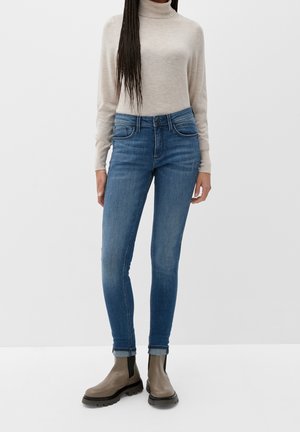 Personne portant un pull à col roulé beige, un jean skinny bleu et des bottines taupe se tient devant un fond uni clair.