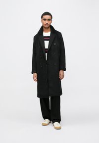 Sort dobbeltradet frakke med stor krave, sat sammen med en creme-stribet sweater, sorte bukser og beige sneakers. Glat stofstruktur.