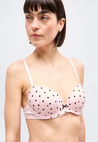 Γυναίκα με κοντά καστανά μαλλιά που φοράει ένα ανοιχτό ροζ δαντελένιο σουτιέν με μαύρα πουά και λεπτές τιράντες, κοιτάζοντας προς τα αριστερά της.