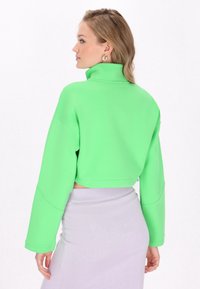 Rövid fazonú, hosszú ujjú pulóver élénk zöld színben, magas gallérral és laza szabással. Világos szürke, texturált szoknyával párosítva.