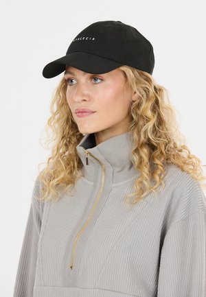 Kvinde med krøllet blondt hår iført en sort baseballkasket og grå ribstrikket pullover med en gylden lynlås, kigger til siden mod en hvid baggrund.
