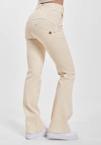 Person med kremfargede flagrende bukser og hvite joggesko, vist bakfra mot en enkel lys bakgrunn.