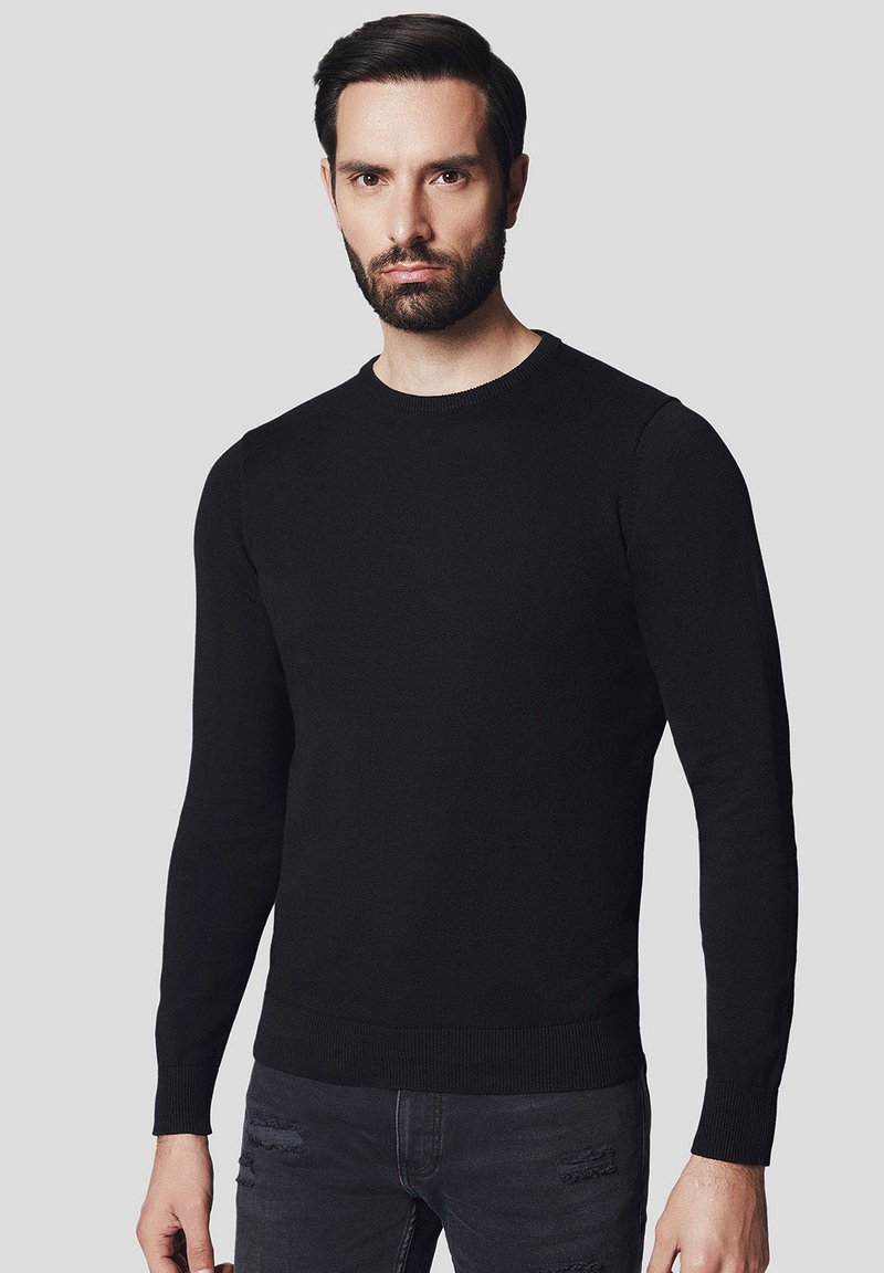 Czarny, długi sweter z okrągłym dekoltem wykonany z gładkiego materiału; posiada ściągacze przy mankietach i dolnej części, noszony z podartymi szarymi jeansami.