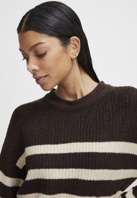 Maglione marrone a coste con strisce crema e marrone scuro, caratterizzato da un collo alto e una texture morbida. Orecchini dorati e argentati visibili.