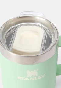 Zaļš nerūsējošā tērauda krūze ar caurspīdīgu vāciņu un baltu silikona blīvējumu. Iespējojusi teksturētu virsmu un izteiktu zīmola logotipu.