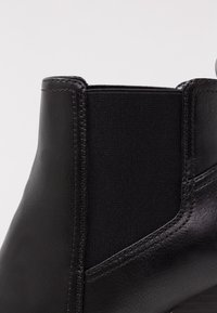 Detailní záběr na černou koženou Chelsea botu s elastickým postranním panelem a výrazným prošíváním na bílém pozadí.