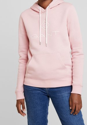 Osoba ubrana w jasnoróżową bluzę z kapturem GANT z białymi sznureczkami i przednią kieszenią, nosząca niebieskie dżinsy, na jednolitym szarym tle.