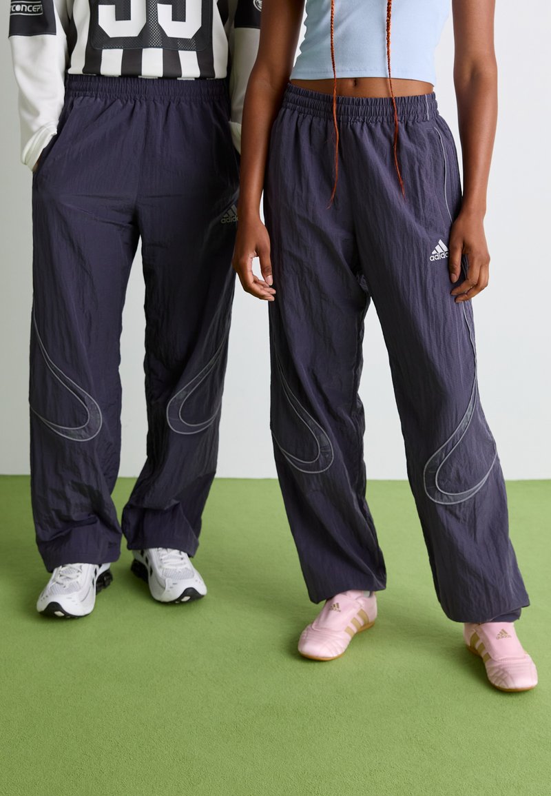 Dvaja ľudia stoja na zelenej podlahe, oblečení v čiernych Adidas teplákových nohaviciach, bielych a ružových športových topánkach; jeden má na sebe crop top a druhý dlhý rukáv.