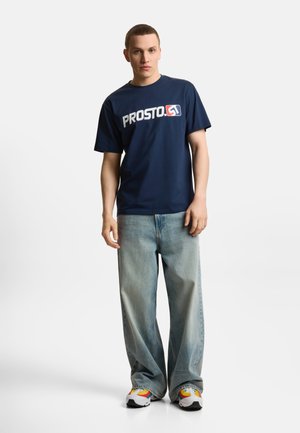 Młody mężczyzna w granatowej koszulce „PROSTO.”, jasnoniebieskich szerokich dżinsach i kolorowych białych trampkach, stojący na białym tle.