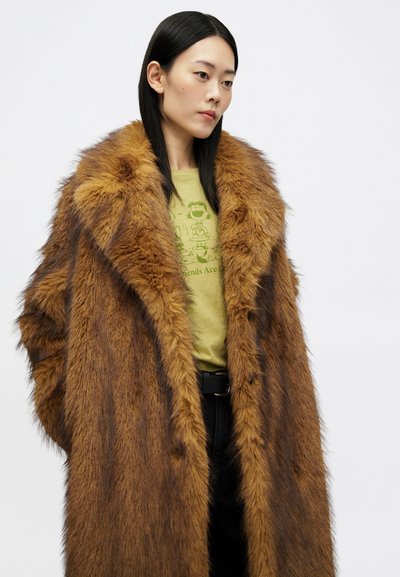 Žena s dlhými čiernymi vlasmi má na sebe prehnane veľký hnedý kožúšok z umelého rúna, svetlozelené tričko s grafikou a čierne nohavice, stojí na bielom pozadí.