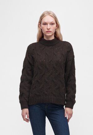 Ung kvinde med langt blondt hår iført en mørkebrun kabelstrikket sweater og blå jeans, stående mod en ensartet lys baggrund.