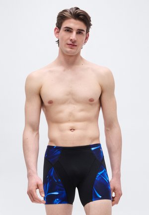 Nuori mies, jolla on lyhyet ruskeat hiukset ja joka on pukeutunut mustiin ja sinisiin uimashortseihin, seisoo yksinkertaisen vaalean taustan edessä.