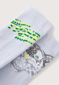 Chaussettes bleu clair présentant un motif du visage d'un tigre en gris, avec des yeux verts et des accents jaunes, accompagnées de texte audacieux en vert.
