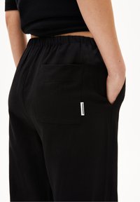 Svarta byxor med elastisk midja, sidoficka och en enda bakficka med en liten varumärkesetikett. Slät textur.