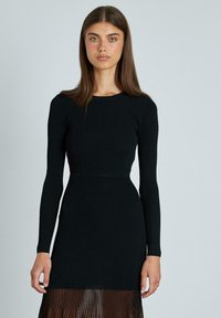 Abito nero a costine con maniche lunghe, corpino aderente e gonna plissettata semi-trasparente. Tessuto liscio, scollo rotondo semplice e design strutturato.