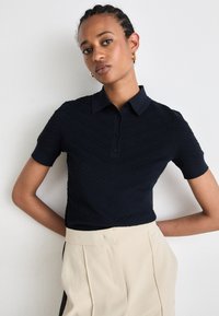 Polo shirt blu navy con maniche corte, caratterizzato da un motivo a zig zag testurizzato, tre bottoni al colletto, abbinato a pantaloni beige.