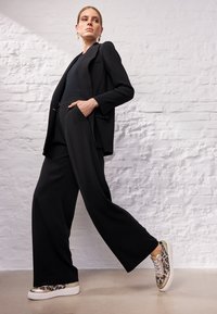 Schwarze, taillierte Blazerjacke über einem eng anliegenden Oberteil, kombiniert mit schwarzen, weit geschnittenen Hosen. Die Sneakers sind mit einem Leopardenmuster und metallischen Akzenten versehen.