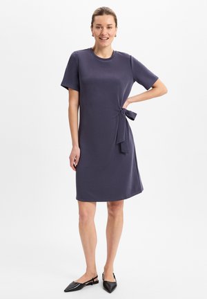 Robe courte bleu marine à manches courtes avec un col rond et un détail de nouage sur le côté. Tissu lisse et confortable avec une coupe ample. Ballerines noires à bout pointu.